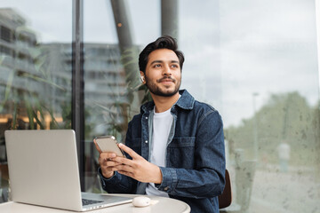 Smiling Indian man, holding mobile phone, looking away, working laptop computer