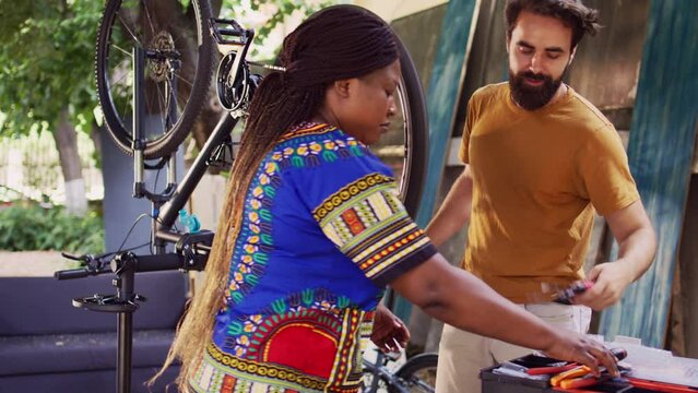 Young sports-loving multiracial couple repairing broken bicycle using professional equipment. Caucasian male loosening and dismantling damaged bike tire while black woman prepares work tools.