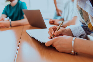 A female hand diligently records essential business ideas in a notebook while engaging in a discussion with colleagues about solving corporate challenges, portraying effective teamwork, communication