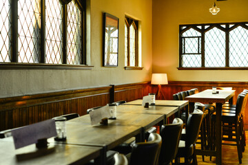The interior of a pub with a row of narrow tables along a wall of windows. There are black wooden chairs on the outside of the tables and a bench along the wall. The walls are pale yellow in color. 