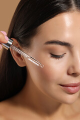 Beautiful young woman applying serum onto her face on beige background, closeup