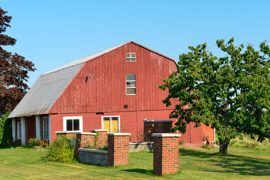 A vintage red two-story barn with white trim, a grey shingled roof with multiple doors and windows. The old farm building is near a large 
with green grass yard and large brick columns as fence posts.
