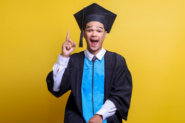 Young Asian male student in mortar board and bachelor gown looking at camera with happy excited expression while pointing finger up 
