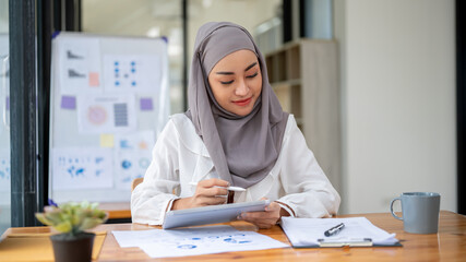 An attractive Asian Muslim businesswoman wearing a hijab focuses on financial reports