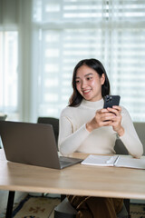 A beautiful Asian woman is looking at her laptop screen and holding her smartphone