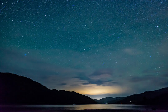 starry sky above the shape of mountains with the light of a city in a fjord in the Marlborough Sounds, New Zealand