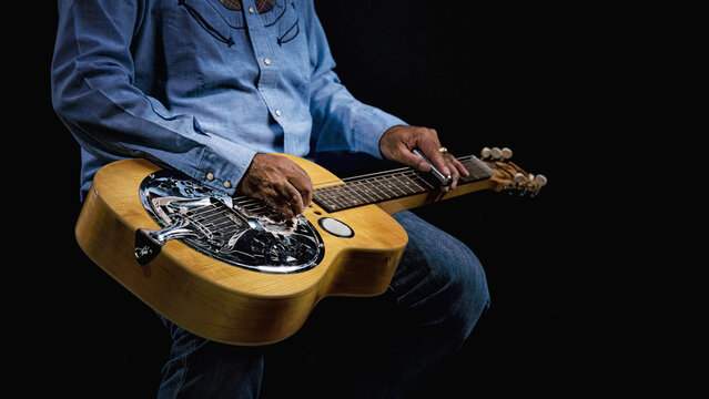 Close-up of a Dobro guitar resting on the lap of a musician dressed in cowboy attire on dark backdrop