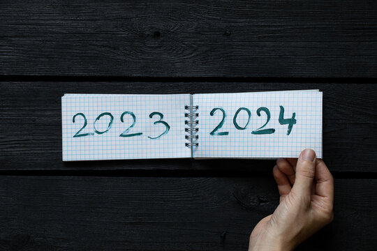 Notepad with the text 2023 and 2024 drawn with paint and a woman's hand turns over a page of a notebook on a black wooden board, new year 2024