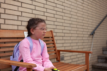 A girl in glasses and with a backpack in a pink sweater sits thoughtfully on a wooden bench near a...