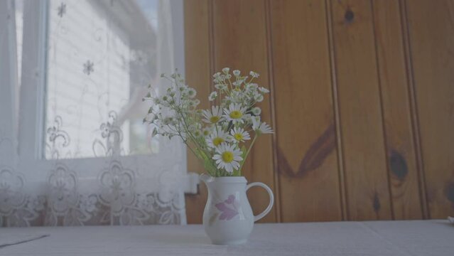 Light vase with bouquet of field daisies stands on table in vintage apartment. Concept of decorative element against window with patterned tulle in country house