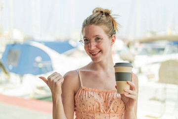 French girl with glasses holding a take away coffee at outdoors pointing to the side to present a product