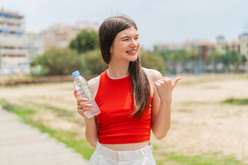 Young pretty Ukrainian woman with a bottle of water at outdoors pointing to the side to present a product