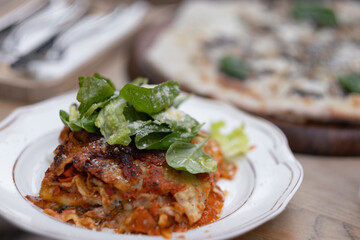 Freshly baked lasagna is served up at an Italian-themed kosher restaurant. Fresh salad greens dress the lasagna. In the background, a pizza and table setting are visible.