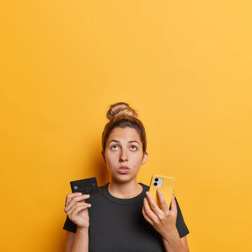 Vertical image of surprised young European woman with hair bun thinking of online shopping holding smartphone with plastic credit card wears casual black t shirt isolated over yellow background