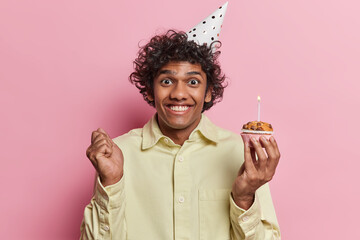 Positive curly haired Hindu man holds small delicious muffin with burning candle clenches fist...