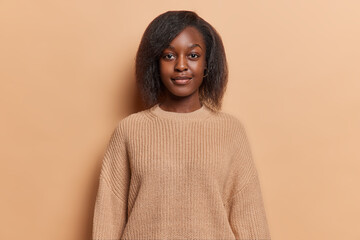Portrait of serious dark skinned woman with short hair full lips and tender expression wears casual knitted jumper focused at camera isolated over brown background. Human facial expressions.