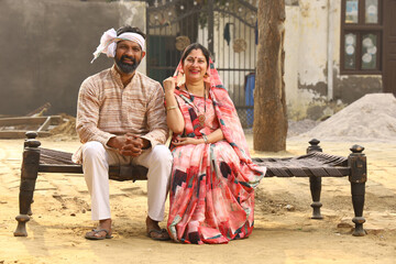 Happy Indian Rural family in village. Husband and wife sitting on cot outside their home front yard. man in kurta pajamas and wife in beautiful saree.
