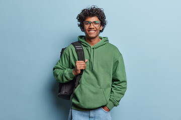 People positive emotions concept. Studio waist up of young happy smiling Hindu male student standing in centre isolated on blue background wearing green hoodie and jeans with black bag on shoulder - obrazy, fototapety, plakaty