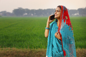 Happy rural Indian woman standing in a mustard agricultural field and surfing through the mobile phone in her hand.