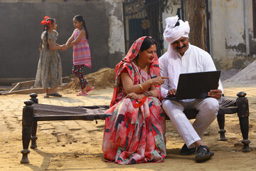 Happy rural Indian husband and wife sitting together outside the cottage / home/ house holding a laptop in hands and surfing online.