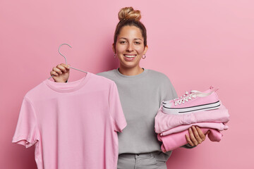 Photo of beautiful cheerful young woman with hairbun holds pink t shirt on hanger folded clothes...