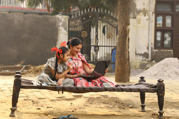 Happy Indian Rural family in village. Rural couple sitting together smiling on cot outside their home in front yard using laptop for online payments.