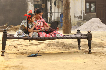 Happy Indian Rural family in village. Rural couple sitting together smiling on cot outside their home in front yard using laptop for online payments.