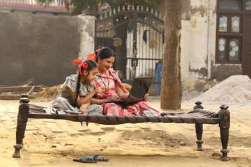 Happy Indian Rural family in village. Rural couple sitting together smiling on cot outside their home in front yard using laptop for online payments.