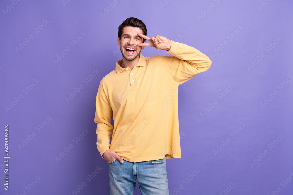 Sticker Photo of excited funky man dressed yellow shirt showing v-sing cover eye isolated purple color background