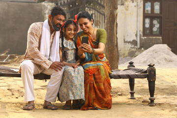 Happy Indian Rural family of Mother, father and daughter sitting together smiling on cot outside their home in front yard using mobile phone.