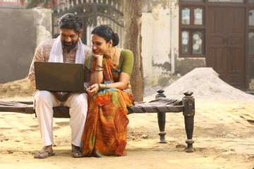 Happy rural Indian husband and wife sitting together outside the cottage / home/ house holding a laptop in hands and surfing online.