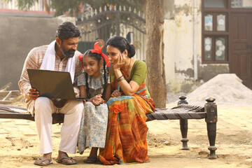 Happy Indian Rural family of Mother, father, daughter sitting on cot outside their home in front yard using Laptop. traditional wear sari kurta pajama