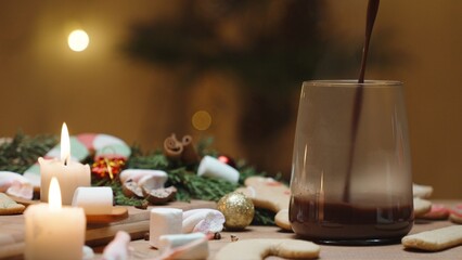 Obraz na płótnie Canvas Christmas table with sweets and candles along with other decorations. Gingerbread cookies are scattered across the table. Hot cocoa is being poured into a mug.