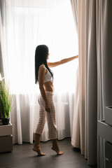 Single woman is a beautiful Caucasian woman in pajamas without standing at the window of a house or in a hotel room and looking at the street on a sunny day