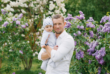 A handsome dad is holding a baby son in his arms, walking through the spring park and smiling. Father's Day