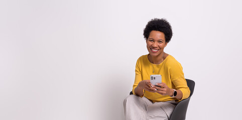 Portrait of young woman smiling and using mobile phone while relaxing on chair over white background