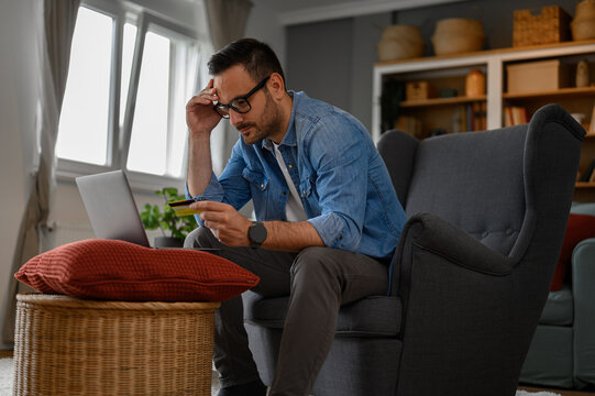 Stressed businessman with credit card paying bills online over laptop while sitting in home office