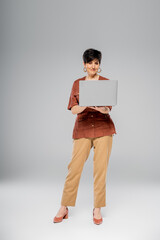 mature businesswoman in brown jacket using laptop on grey, autumn, seasonal attire, fashion trend