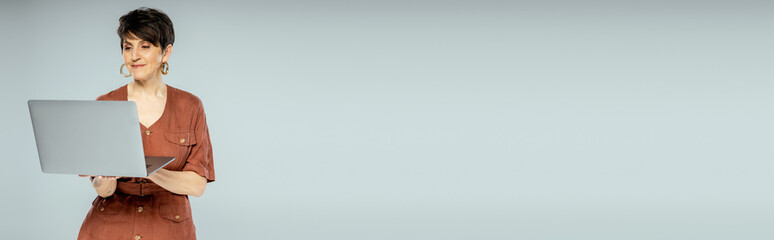 brunette businesswoman in brown jacket using laptop on grey, autumn fashion, stylish look, banner