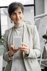 smiling middle aged businesswoman in earphones holding smartphone and looking away in office