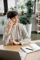 successful middle aged corporate manager talking on smartphone near laptop and notebook in office