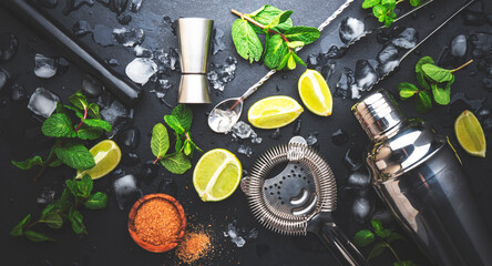 Mojito or caipirinha cocktail preparation. Mint, cane sugar, lime, ice and steel bar tools: shaker, jigger, strainer, muddler on dark stone background, top view