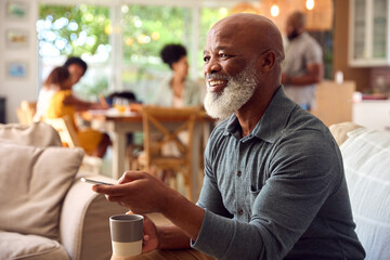 Senior Man Sitting On Sofa At Home Watching TV With Multi-Generation Family In Background