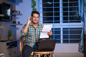 Technology, remote job and lifestyle concept - happy Indian man with laptop computer working at home office and talking on mobile phone while checking papers.