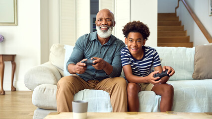 Grandfather And Grandson On Sofa At Home Playing Video Game Together With Boy Celebrating Win