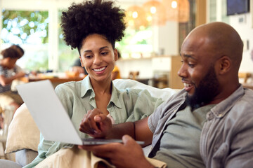 Couple At Home Using Laptop To Shop Or Book Holiday With Multi-Generation Family In Background