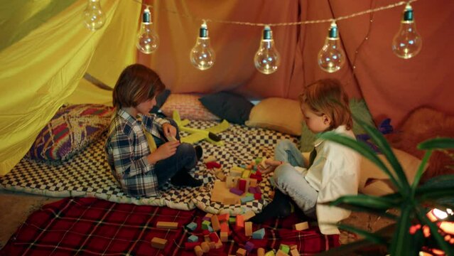 Two young boys sitting cross legged inside a spacious indoor sheet fort while helping each other out with building blocks enjoying each others company