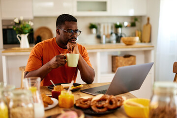 Fototapeta na wymiar Pensive black man working on laptop during breakfast at home
