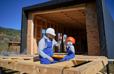 Father with toddler son building wooden frame house. Male builder giving high five to kid on construction site, wearing helmet and blue overalls on sunny day. Carpentry and family concept.