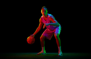 Dynamic image of teen boy in uniform, playing basketball against black studio background in neon light. Dribbling ball. Concept of professional sport, competition, hobby, game, competition